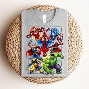May include: A grey t-shirt with a graphic of Marvel superheroes. The design includes Iron Man, Captain America, Spider-Man, Thor, Wolverine, Hulk, and Deadpool. The superheroes are depicted in action poses with bright colours and detailed illustrations.