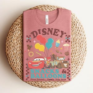 May include: A dusty rose t-shirt featuring a graphic design with the word "DISNEY" in brown, along with cartoon cars, balloons, and snacks. The text "SNACKS & BALLOONS" is below the Disney logo, with "Pixar Friends &bull; Family Trip 2026" at the bottom.