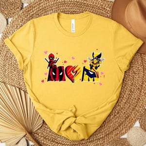 Op de afbeelding: Mosterdgele t-shirt met een afbeelding van Deadpool en Wolverine. Het ontwerp bevat het woord "Love" met hartaccenten. Het t-shirt is gemaakt van zacht materiaal en heeft een klassieke ronde hals.