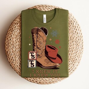 May include: An olive green t-shirt featuring a graphic of a brown cowboy boot, a brown cowboy hat, and two stacked blocks with the text T5. Below the graphic, the text reads I KNEW IT I KNEW YOU. The design includes small star and flower icons.
