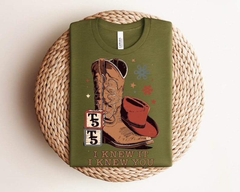 May include: An olive green t-shirt featuring a graphic of a brown cowboy boot, a brown cowboy hat, and two stacked blocks with the text T5. Below the graphic, the text reads I KNEW IT I KNEW YOU. The design includes small star and flower icons.