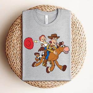 May include: A heather gray t-shirt featuring a colorful graphic of Woody and Jessie from Toy Story riding a horse. The design includes a red lasso and the characters in their signature outfits. The shirt is laid on a woven surface.