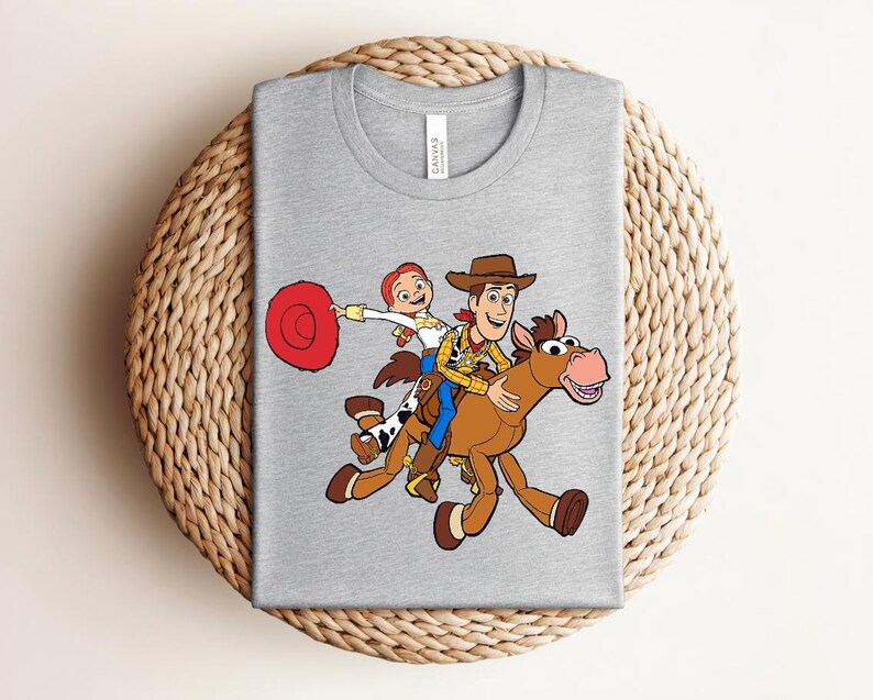 May include: A heather gray t-shirt featuring a colorful graphic of Woody and Jessie from Toy Story riding a horse. The design includes a red lasso and the characters in their signature outfits. The shirt is laid on a woven surface.