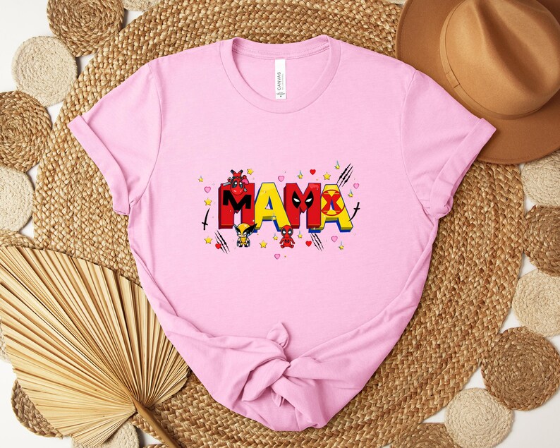 Puede incluir: Camiseta rosa claro con la palabra "MAMA" en letras coloridas estilo c&oacute;mic. El dise&ntilde;o incluye personajes y s&iacute;mbolos de dibujos animados, con corazones y estrellas. La camiseta est&aacute; doblada.