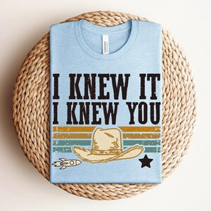 May include: A light blue t-shirt featuring the black text I KNEW IT I KNEW YOU above a graphic of a beige cowboy hat, a small rocket, and a black star, set against horizontal stripes in muted yellow, green, and blue colors with a distressed finish.