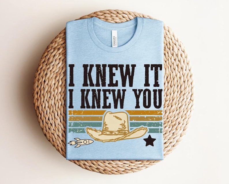 May include: A light blue t-shirt featuring the black text I KNEW IT I KNEW YOU above a graphic of a beige cowboy hat, a small rocket, and a black star, set against horizontal stripes in muted yellow, green, and blue colors with a distressed finish.