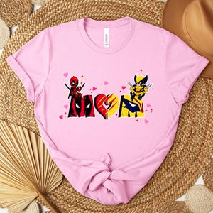Op de afbeelding: Lichtroze t-shirt met een grafisch ontwerp met het woord "LOVE" in een hartvorm, met Deadpool- en Wolverine-personages. Het ontwerp bevat rode, zwarte, gele en blauwe kleuren, met kleine roze hartjes.
