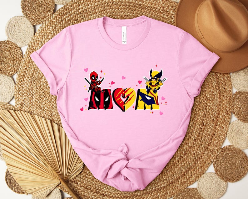 Op de afbeelding: Lichtroze t-shirt met een grafisch ontwerp met het woord "LOVE" in een hartvorm, met Deadpool- en Wolverine-personages. Het ontwerp bevat rode, zwarte, gele en blauwe kleuren, met kleine roze hartjes.