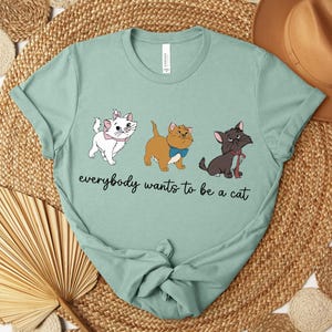 May include: A light green t-shirt featuring three cartoon cats with the text "everybody wants to be a cat". The cats are white, brown, and black, each with unique accessories. The shirt is made of a soft material.