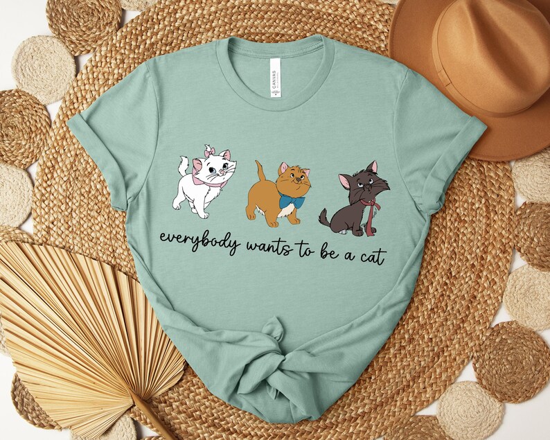 May include: A light green t-shirt featuring three cartoon cats with the text "everybody wants to be a cat". The cats are white, brown, and black, each with unique accessories. The shirt is made of a soft material.