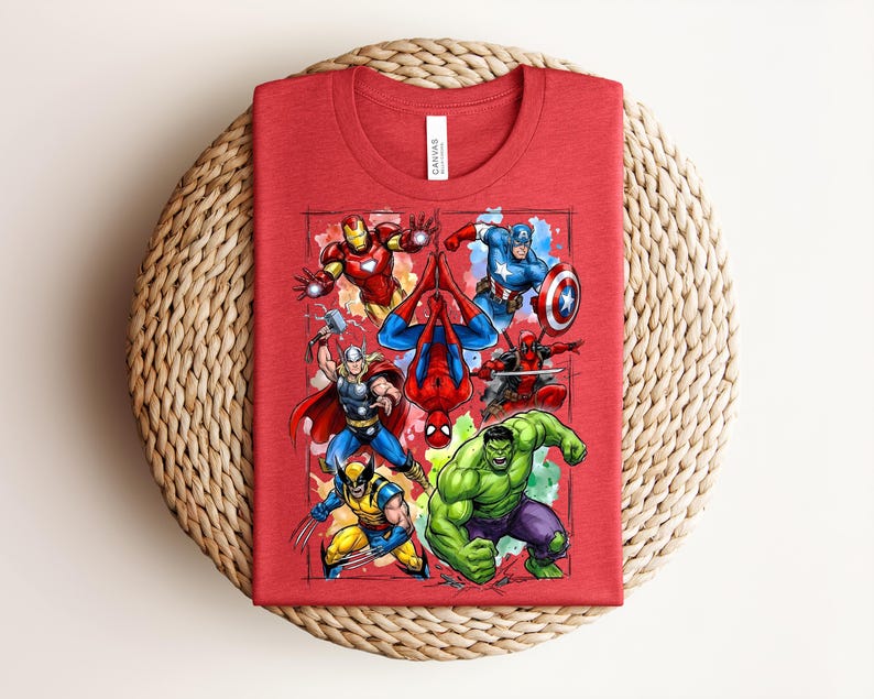May include: Red t-shirt with a vibrant graphic of Marvel superheroes, such as Iron Man, Spider-Man, Captain America, Thor, Wolverine, Hulk, and Deadpool. The folded shirt is presented on a woven, circular mat.