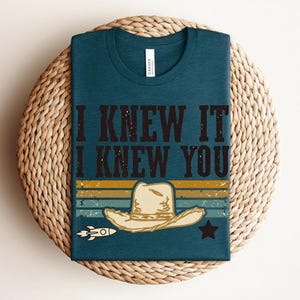 May include: A dark teal crew neck t-shirt featuring the text I KNEW IT I KNEW YOU in a distressed black font. Below the text is a graphic of a beige cowboy hat, a small white rocket, and a black star set against horizontal stripes.