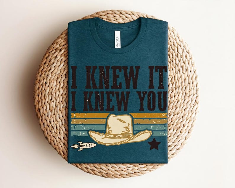 May include: A dark teal crew neck t-shirt featuring the text I KNEW IT I KNEW YOU in a distressed black font. Below the text is a graphic of a beige cowboy hat, a small white rocket, and a black star set against horizontal stripes.