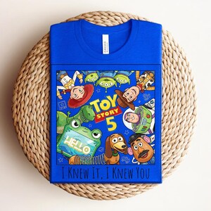K&ouml;nnte beinhalten: Ein k&ouml;nigsblaues T-Shirt mit einem Toy Story 5-Grafikdruck, der Charaktere wie Woody, Buzz Lightyear, Jessie, Rex, Slinky Dog, Mr. Potato Head und Aliens zeigt. Das Design enth&auml;lt den Text Toy Story 5 und I Knew It, I Knew You.