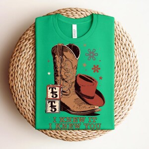 May include: A bright green t-shirt featuring a brown cowboy boot, a brown cowboy hat, and two stacked blocks with the text T5. The graphic includes small star and flower shapes in red and brown. The text I KNEW IT I KNEW YOU is printed below.