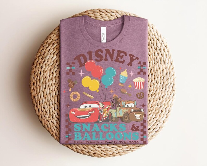May include: A purple t-shirt featuring a graphic design with the text "Disney Snacks & Balloons". The design includes cartoon cars, balloons, and food icons. The shirt is on a woven, circular surface.