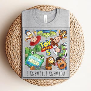 K&ouml;nnte beinhalten: Ein graues T-Shirt mit einem Grafikdruck von Toy Story-Figuren, darunter Woody, Buzz Lightyear, Jessie, Rex, Slinky Dog, Mr. Potato Head und Aliens. Der Text lautet Toy Story 5 und I KNEW IT, I KNEW YOU.