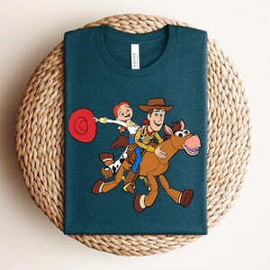 May include: Teal crew neck t-shirt featuring a colorful cartoon illustration of Woody, Jessie, and Bullseye from Toy Story. The design shows the characters riding on Bullseye. The shirt has a tag that says "Canvas".