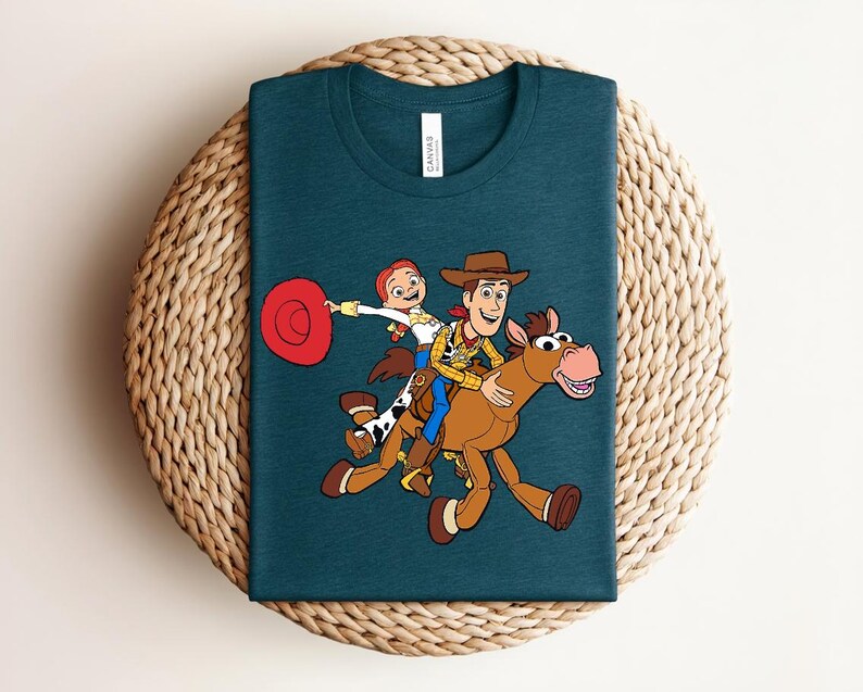 May include: Teal crew neck t-shirt featuring a colorful cartoon illustration of Woody, Jessie, and Bullseye from Toy Story. The design shows the characters riding on Bullseye. The shirt has a tag that says "Canvas".
