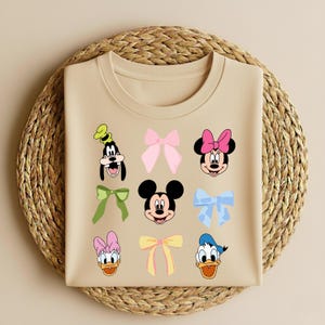 May include: A beige t-shirt with cartoon character faces and bows. Characters include Goofy, Minnie Mouse, Mickey Mouse, Donald Duck, and Daisy Duck. Bows are pink, green, yellow, and blue.