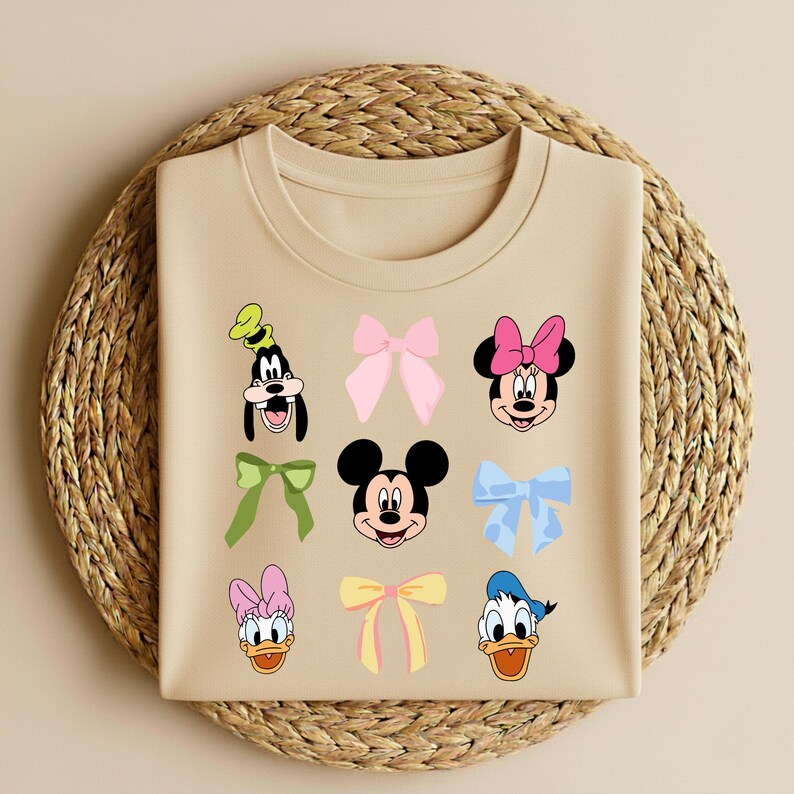 May include: A beige t-shirt with cartoon character faces and bows. Characters include Goofy, Minnie Mouse, Mickey Mouse, Donald Duck, and Daisy Duck. Bows are pink, green, yellow, and blue.