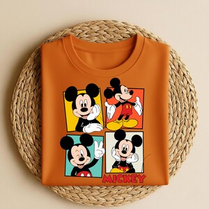 以下が含まれることがあります： オレンジ色のクルーネックTシャツ。ミッキーマウスが様々なポーズをとる4つのパネルグラフィックが特徴です。各パネルは異なる背景色です。「MICKEY」の文字が赤色で下部にプリントされています。Tシャツは織りマットの上に折りたたまれています。