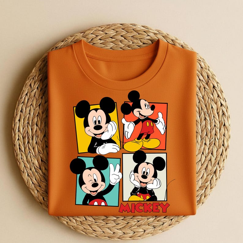以下が含まれることがあります： オレンジ色のクルーネックTシャツ。ミッキーマウスが様々なポーズをとる4つのパネルグラフィックが特徴です。各パネルは異なる背景色です。「MICKEY」の文字が赤色で下部にプリントされています。Tシャツは織りマットの上に折りたたまれています。