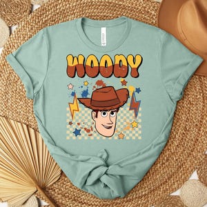 Puede incluir: Camiseta de color verde azulado claro con la palabra "WOODY" en fuente retro marr&oacute;n y amarilla. La camiseta tiene un gr&aacute;fico de un vaquero de dibujos animados con un sombrero marr&oacute;n, estrellas y rayos.