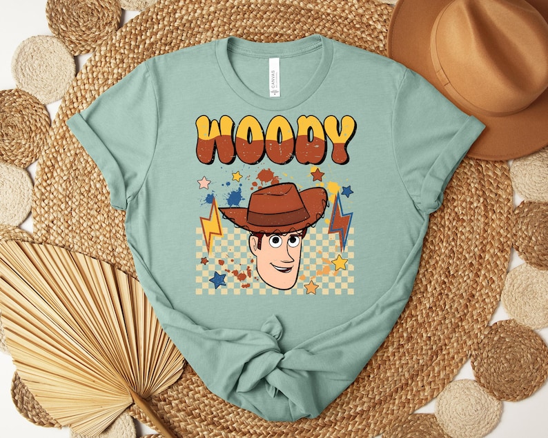Puede incluir: Camiseta de color verde azulado claro con la palabra "WOODY" en fuente retro marr&oacute;n y amarilla. La camiseta tiene un gr&aacute;fico de un vaquero de dibujos animados con un sombrero marr&oacute;n, estrellas y rayos.