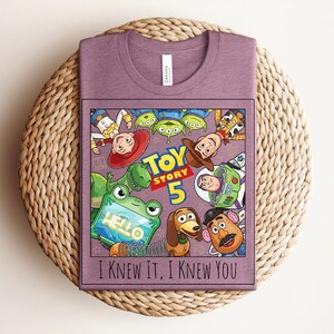 K&ouml;nnte beinhalten: Ein malvenfarbenes T-Shirt mit Rundhalsausschnitt und einem Grafikdruck von Toy Story-Figuren, darunter Woody, Buzz Lightyear, Jessie, Rex, Slinky Dog, Mr. Potato Head und Aliens. Der Text lautet Toy Story 5 und I KNEW IT, I KNEW YOU.