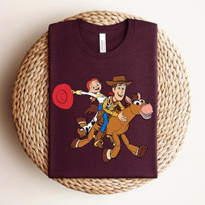May include: A maroon t-shirt featuring a colorful cartoon illustration of Woody, Jessie, and a horse from the Toy Story movie. The characters are riding the horse. The shirt has a tag on the collar.