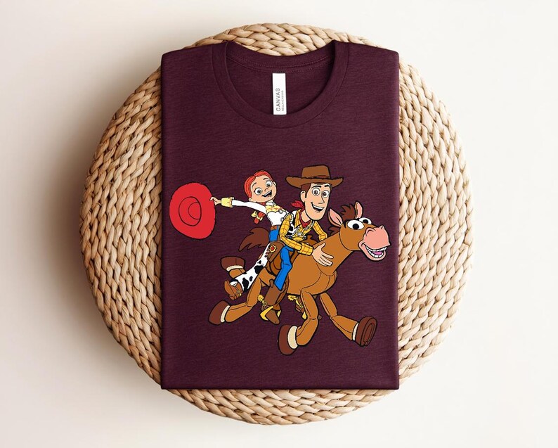 May include: A maroon t-shirt featuring a colorful cartoon illustration of Woody, Jessie, and a horse from the Toy Story movie. The characters are riding the horse. The shirt has a tag on the collar.