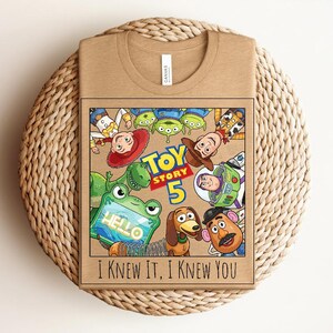 K&ouml;nnte beinhalten: Ein beigefarbenes T-Shirt mit einem Grafikdruck von Toy Story-Figuren, darunter Woody, Buzz Lightyear, Jessie, Rex, Slinky Dog, Mr. Potato Head und Aliens. Der Text lautet Toy Story 5 und I Knew It, I Knew You mit einem Frosch, der ein Tablet h&auml;lt.