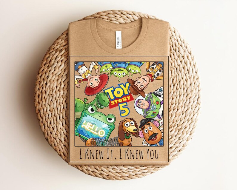 K&ouml;nnte beinhalten: Ein beigefarbenes T-Shirt mit einem Grafikdruck von Toy Story-Figuren, darunter Woody, Buzz Lightyear, Jessie, Rex, Slinky Dog, Mr. Potato Head und Aliens. Der Text lautet Toy Story 5 und I Knew It, I Knew You mit einem Frosch, der ein Tablet h&auml;lt.