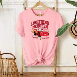 May include: Pink t-shirt with a red cartoon race car graphic and the text "Lightning McQueen" in red. The car is driving with a trophy and the text "Piston Cup Racing Series".