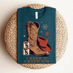 May include: A dark teal t-shirt featuring a graphic of a brown cowboy boot, a brown cowboy hat, and two stacked alphabet blocks with the letter T and the number 5. The text "I KNEW IT I KNEW YOU" is printed below the graphic with small star and flower icons.