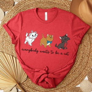 May include: Red t-shirt featuring three cartoon cats with the text "everybody wants to be a cat". The cats are white, brown, and gray, each with unique accessories. The shirt is knotted at the bottom.
