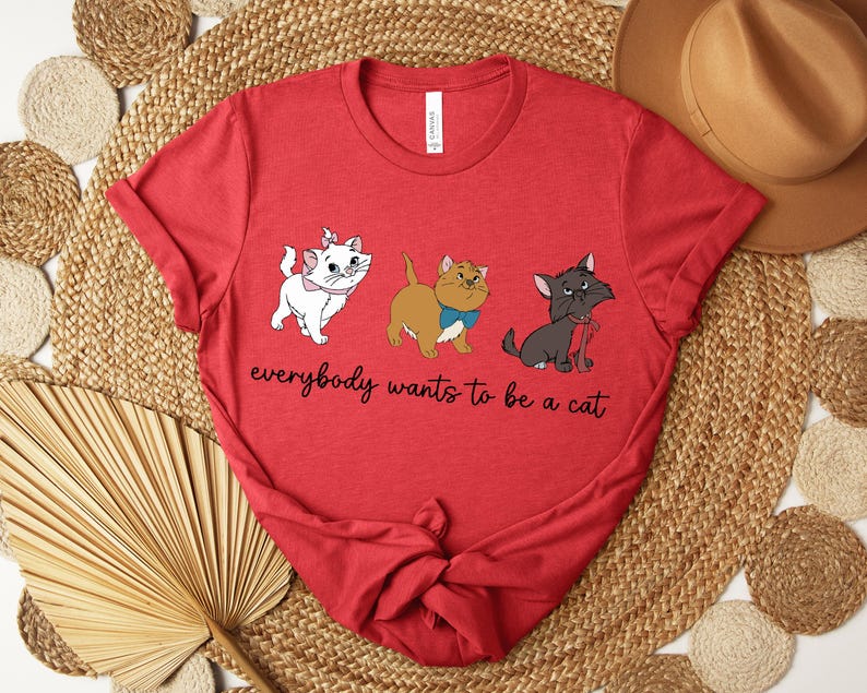 May include: Red t-shirt featuring three cartoon cats with the text "everybody wants to be a cat". The cats are white, brown, and gray, each with unique accessories. The shirt is knotted at the bottom.