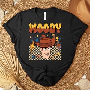 Puede incluir: Camiseta negra con una ilustraci&oacute;n de dibujos animados de Woody de Toy Story. El dise&ntilde;o incluye el nombre "WOODY" en letras naranjas y marrones, un sombrero de vaquero y un fondo a cuadros. Tambi&eacute;n se incluyen estrellas y rayos.