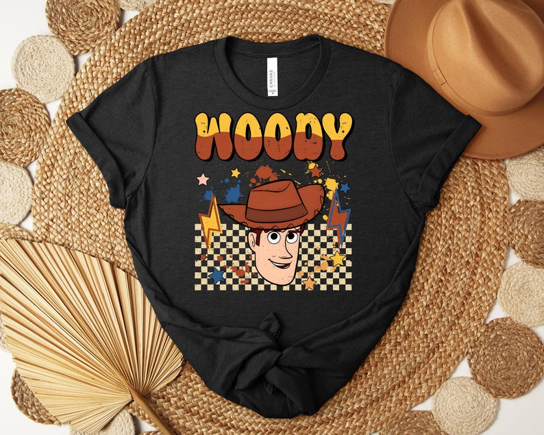 Puede incluir: Camiseta negra con una ilustraci&oacute;n de dibujos animados de Woody de Toy Story. El dise&ntilde;o incluye el nombre "WOODY" en letras naranjas y marrones, un sombrero de vaquero y un fondo a cuadros. Tambi&eacute;n se incluyen estrellas y rayos.