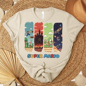 May include: Beige t-shirt featuring a colorful Super Mario graphic. The design includes four vertical panels with scenes from the game, including Mario, a castle, Yoshi, and space. The words "SUPER MARIO" are printed in blue and orange.