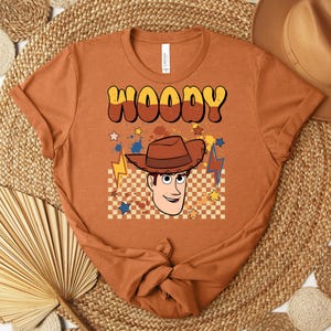 Puede incluir: Una camiseta naranja con la palabra "WOODY" en una fuente retro, y una imagen de dibujos animados de un vaquero. El dise&ntilde;o incluye un sombrero de vaquero, estrellas y rayos. La camiseta est&aacute; anudada en la parte inferior.