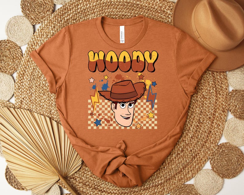 Puede incluir: Una camiseta naranja con la palabra "WOODY" en una fuente retro, y una imagen de dibujos animados de un vaquero. El dise&ntilde;o incluye un sombrero de vaquero, estrellas y rayos. La camiseta est&aacute; anudada en la parte inferior.