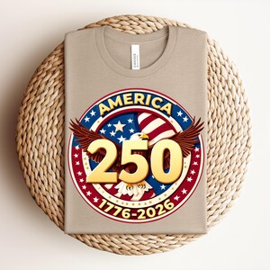 May include: A beige t-shirt with a circular graphic celebrating America's 250th anniversary. The design includes an eagle, stars, and the text "AMERICA 250 1776-2026" in red, white, and blue colours.