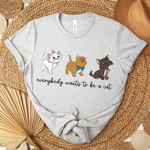 May include: Light gray t-shirt featuring three cartoon cats with the text "everybody wants to be a cat". The cats are white, tan, and black, each with unique accessories. The shirt is made of a soft material.