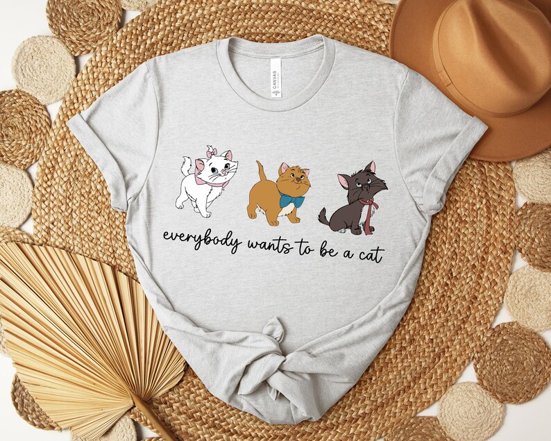 May include: Light gray t-shirt featuring three cartoon cats with the text "everybody wants to be a cat". The cats are white, tan, and black, each with unique accessories. The shirt is made of a soft material.