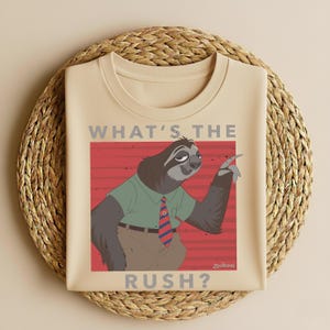 Puede incluir: Camiseta beige con un personaje de perezoso de dibujos animados de Zootopia, que lleva una camisa verde y una corbata a rayas rojas y azules. El texto "WHAT'S THE RUSH?" est&aacute; impreso encima y debajo de la ilustraci&oacute;n del perezoso.