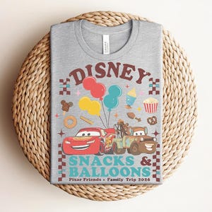 May include: A gray t-shirt featuring a colorful graphic design with the text "Disney Snacks & Balloons". The design includes cartoon cars, balloons, and food icons. The shirt is a crew neck and is made of a soft material.