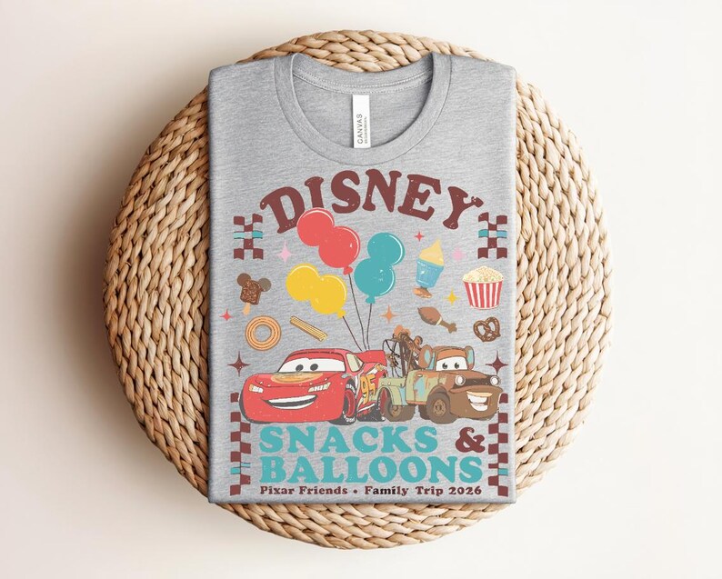 May include: A gray t-shirt featuring a colorful graphic design with the text "Disney Snacks & Balloons". The design includes cartoon cars, balloons, and food icons. The shirt is a crew neck and is made of a soft material.