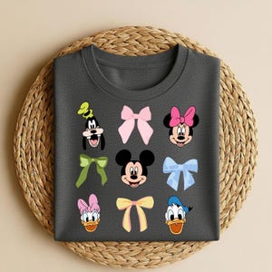 May include: Dark grey t-shirt with cartoon character faces and bows. The design includes Mickey Mouse, Minnie Mouse, Donald Duck, Daisy Duck, and Goofy. The bows are in various colours, including pink, yellow, and blue.